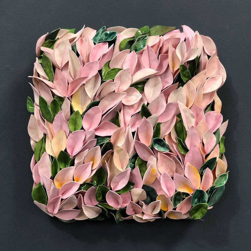 A square wall sculpture featuring layered ceramic leaves in shades of pink, green, and yellow against a dark background.